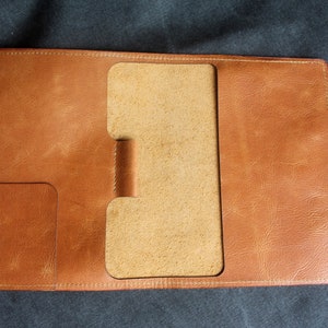 Puede incluir: Una cubierta de cuaderno de cuero marrón con una solapa y un portaplumas. La cubierta tiene un borde cosido y un acabado liso.