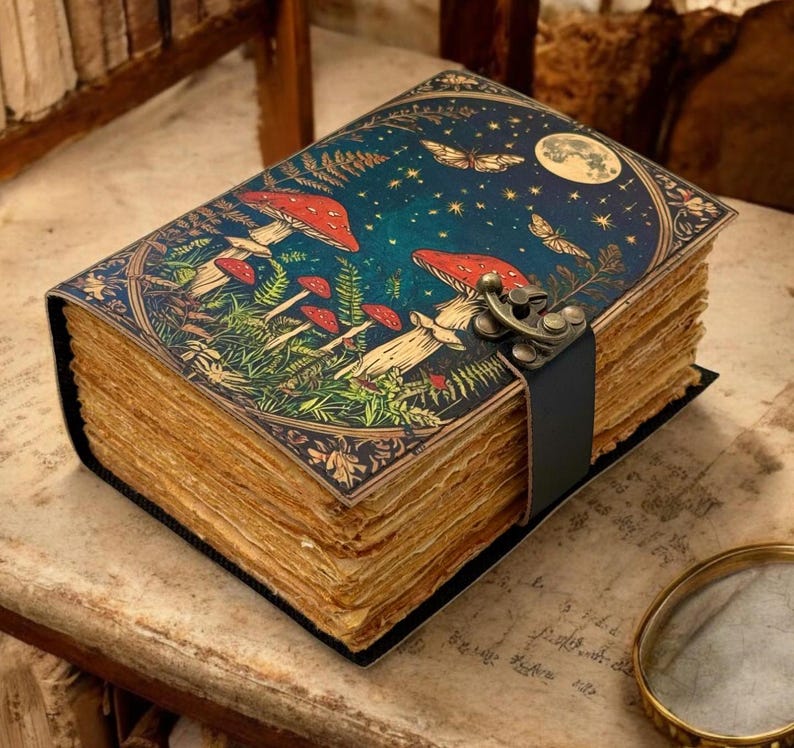 May include: A leather-bound journal with a dark blue cover featuring a whimsical illustration of mushrooms, ferns, and a crescent moon with stars. The journal has a leather strap closure with a metal clasp.