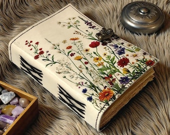 May include: A cream-colored journal with a floral design featuring red, yellow, orange, and purple flowers. The journal has a metal clasp and black stitching along the spine. A small, silver container sits nearby.