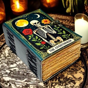 May include: A decorative journal with a skeleton illustration on the cover, featuring a full moon, roses, and the text "THE OVERTHINKER." The book has a teal cover and aged, yellowed pages, with a metal clasp.