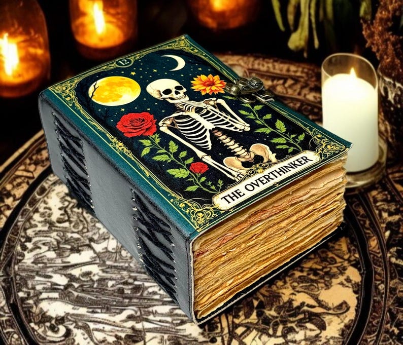 May include: A decorative journal with a skeleton illustration on the cover, featuring a full moon, roses, and the text "THE OVERTHINKER." The book has a teal cover and aged, yellowed pages, with a metal clasp.