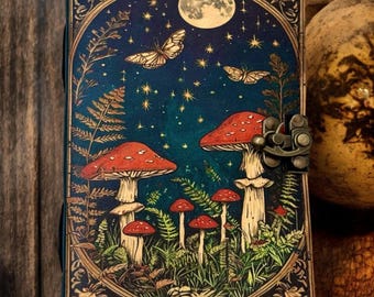 May include: A dark blue leather journal with a colorful illustration of mushrooms, butterflies, and a full moon in a forest setting. The journal has a metal clasp closure.