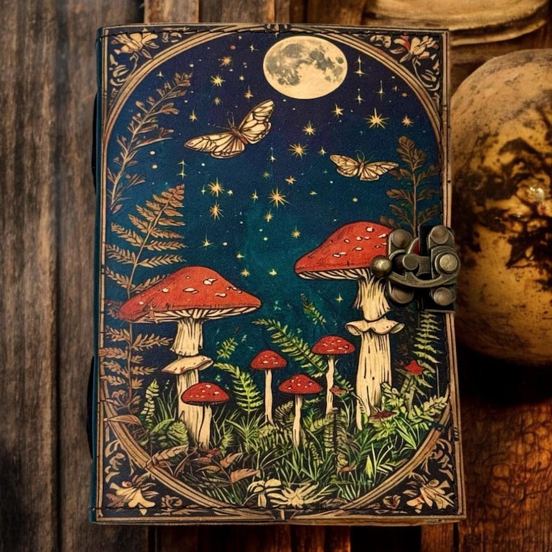 May include: A dark blue leather journal with a colorful illustration of mushrooms, butterflies, and a full moon in a forest setting. The journal has a metal clasp closure.