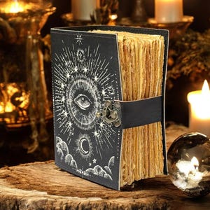 Puede incluir: Un diario negro con un dise&ntilde;o de ojo y celestial, asegurado por una correa y un cierre negros. Las p&aacute;ginas envejecidas son visibles. El diario descansa sobre una superficie de madera. Velas y una bola de cristal en el fondo.