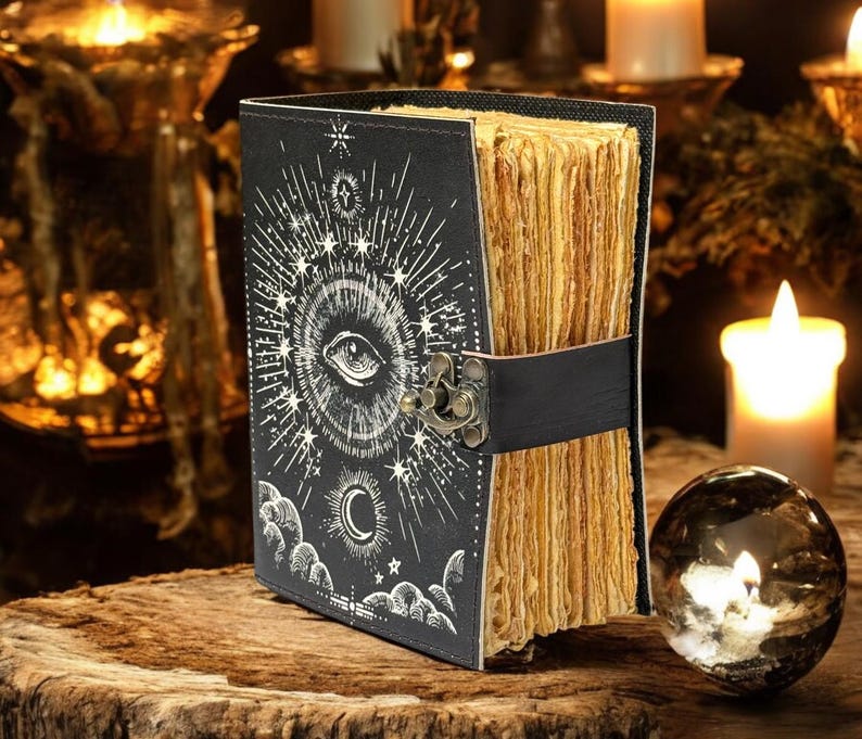 Puede incluir: Un diario negro con un dise&ntilde;o de ojo y celestial, asegurado por una correa y un cierre negros. Las p&aacute;ginas envejecidas son visibles. El diario descansa sobre una superficie de madera. Velas y una bola de cristal en el fondo.