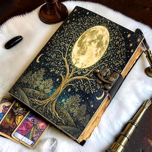 May include: A rectangular journal with a dark blue cover featuring a large yellow full moon behind a tree with golden branches and leaves. The journal has a brass metal latch closure and deckled paper edges, accompanied by three tarot cards.