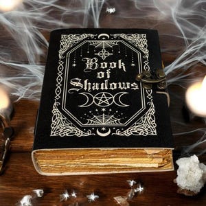 Może przedstawiać: Czarny dziennik "Book of Shadows" z ozdobnymi białymi detalami i metalową klamrą. Okładka zawiera tytuł gotycką czcionką oraz symbole niebieskie. Widoczne są postarzone strony, sugerujące estetykę vintage lub antyczną.