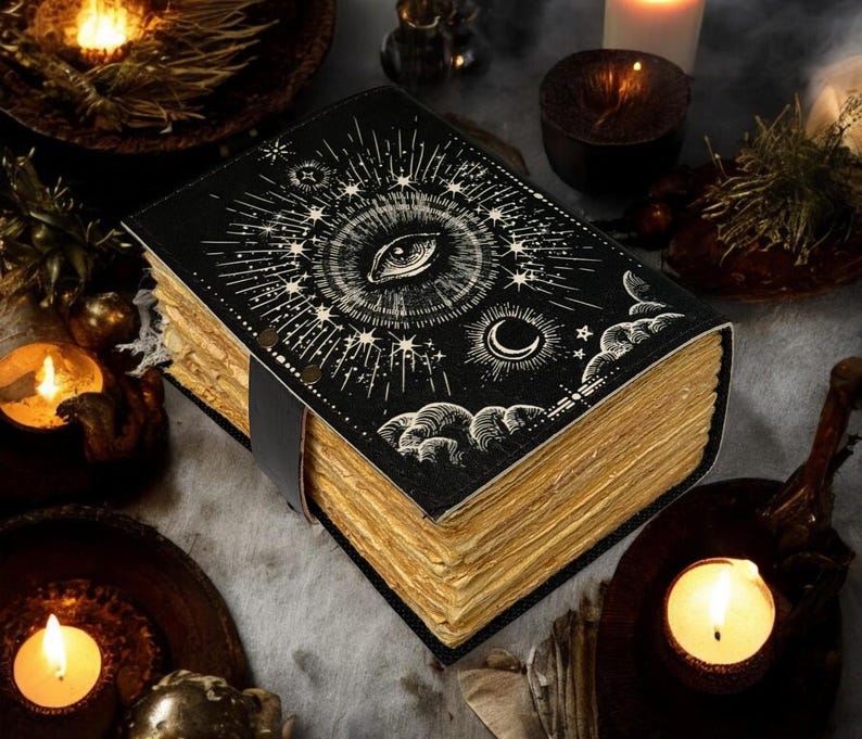 Puede incluir: Un diario de cuero negro con un dise&ntilde;o de ojo y celestial. Las p&aacute;ginas tienen una apariencia vintage y envejecida. Una correa de cuero asegura el diario. La imagen est&aacute; rodeada de velas encendidas y otra decoraci&oacute;n m&iacute;stica.