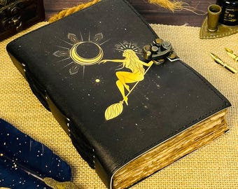 May include: A black leather-bound journal with a gold design of a woman riding a broom under a crescent moon. The journal has a brass clasp and is closed.