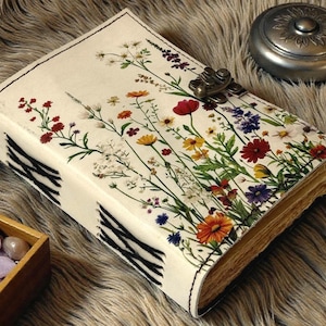 May include: A cream-colored journal with a floral design featuring red, yellow, orange, and purple flowers. The journal has a metal clasp and black and white striped accents on the spine. A small silver container sits nearby.