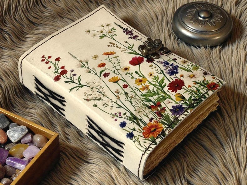 May include: A cream-colored journal with a floral design featuring red, yellow, orange, and purple flowers. The journal has a metal clasp and black and white striped accents on the spine. A small silver container sits nearby.