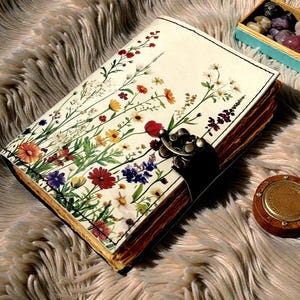 May include: A journal with a floral design on the cover, featuring red, yellow, purple, and white flowers. The journal has a metal clasp and sits on a fluffy, light brown surface. A small wooden box with stones and a wooden container are also present.