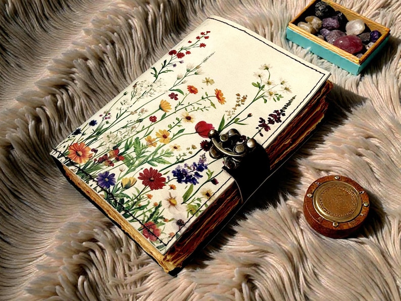 May include: A journal with a floral design on the cover, featuring red, yellow, purple, and white flowers. The journal has a metal clasp and sits on a fluffy, light brown surface. A small wooden box with stones and a wooden container are also present.