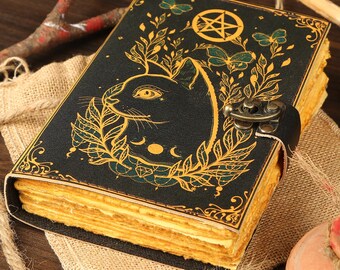 May include: A black leather-bound journal with a gold pentagram and a black cat illustration. The journal has a brass clasp and a leather strap.