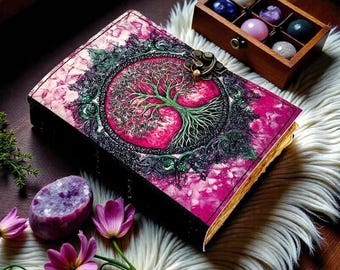 Könnte beinhalten: Ein dekoratives Notizbuch mit einem rosa und grünen Lebensbaum-Design auf dem Einband. Das Notizbuch ist mit einem Metallverschluss verschlossen. Das Bild zeigt auch eine Holzkiste mit bunten Steinen, lila Blumen und einem großen lila Kristall.