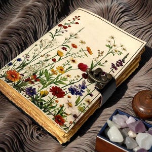 May include: A floral journal with a cream-colored cover featuring colorful wildflowers. The journal has a metal clasp and a black leather strap. The pages have a gold-colored edge. A small wooden box with gemstones and a wooden container are next to the journal.