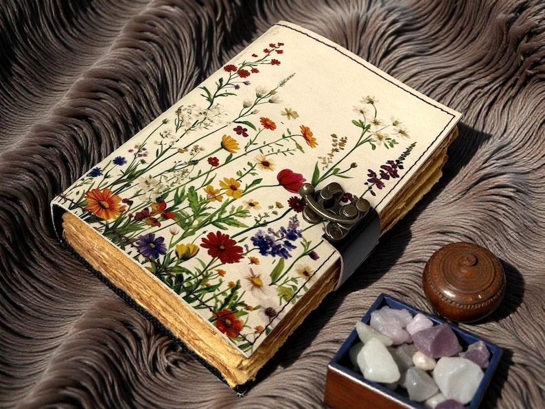 May include: A floral journal with a cream-colored cover featuring colorful wildflowers. The journal has a metal clasp and a black leather strap. The pages have a gold-colored edge. A small wooden box with gemstones and a wooden container are next to the journal.