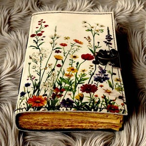 May include: A journal with a floral design cover featuring a variety of colorful wildflowers. The journal has a clasp closure and aged-looking pages. The cover is cream-colored with a black border.