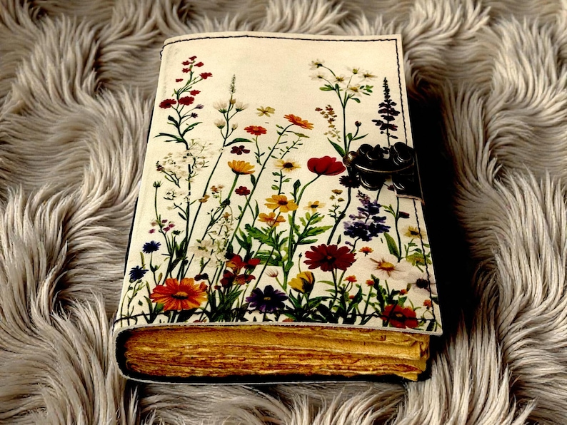May include: A journal with a floral design cover featuring a variety of colorful wildflowers. The journal has a clasp closure and aged-looking pages. The cover is cream-colored with a black border.