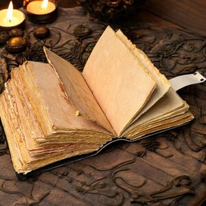 May include: An open, antique-style book with aged, yellowed pages. The book is bound in black and has a white ribbon bookmark. The pages have a rough, uneven edge. The book is on a carved wooden surface, with lit candles in the background.