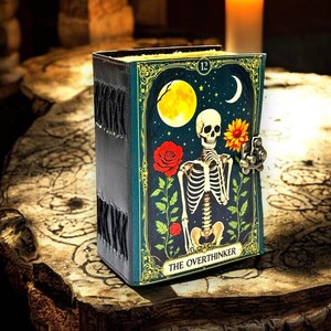 May include: A dark teal journal featuring a skeleton illustration, a full moon, and a red rose. The journal has a metal clasp and the words "THE OVERTHINKER". The spine is black with decorative stitching.
