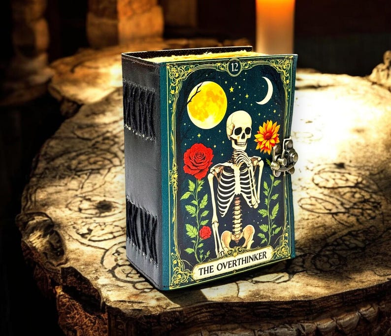 May include: A dark teal journal featuring a skeleton illustration, a full moon, and a red rose. The journal has a metal clasp and the words "THE OVERTHINKER". The spine is black with decorative stitching.
