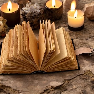 May include: An open, antique-looking journal with brown leather covers and aged, yellowed pages. The journal is lying open on a rustic, brown stone surface. There are several lit candles in the background.