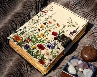 May include: A floral journal with a cream-colored cover adorned with colorful wildflowers. The journal has a metal clasp and a black leather strap. The pages have a vintage, aged appearance. A small wooden box with stones and a wooden container are next to the journal.