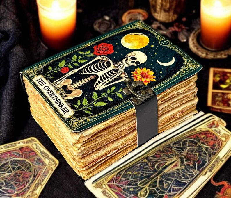 May include: A deck of aged pages bound with a black strap, featuring a decorative cover with a skeleton, flowers, and the text "THE OVERTHINKER." The image includes lit candles and tarot cards, suggesting a mystical theme.