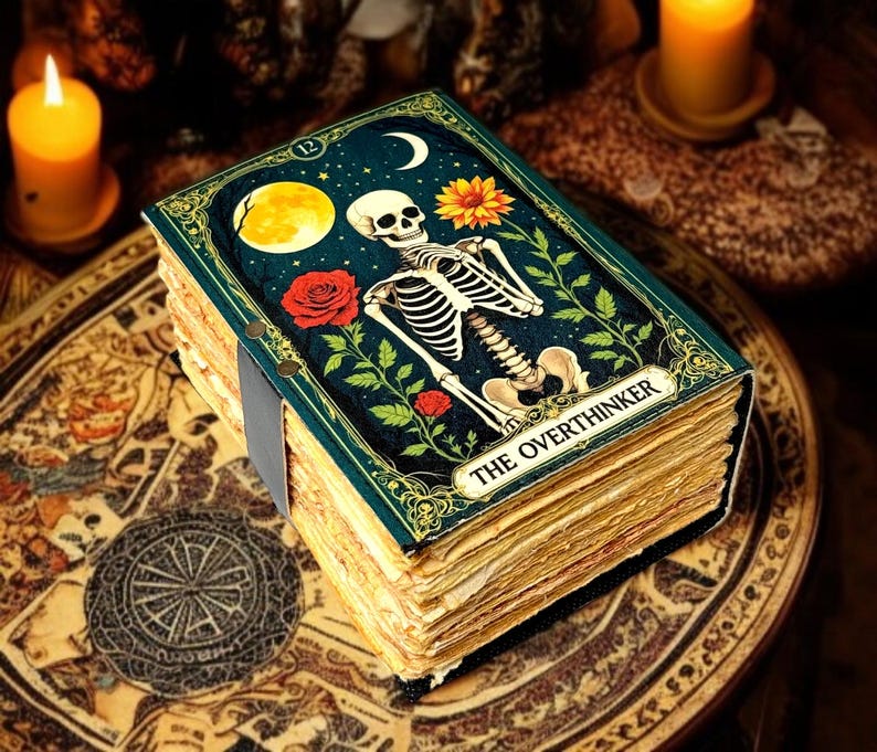 May include: A decorative journal with a dark teal cover featuring a skeleton, moon, and floral elements. The journal's title, "THE OVERTHINKER," is displayed on the cover. The pages are aged, and the journal is bound with a grey ribbon.