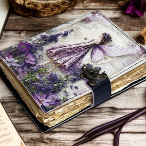 Könnte beinhalten: Ein handgefertigtes Notizbuch mit einer Feenillustration auf dem Einband, umgeben von lila Blumen. Das Notizbuch hat einen schwarzen Lederriemen und Verschluss sowie gealterte Seiten. Das Cover zeigt eine Fee in einem lila Kleid.