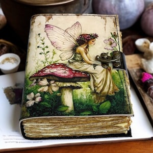 May include: A leather-bound journal with a fantasy illustration. The cover features a fairy sitting on a mushroom, with a red cap and white stem. The fairy has wings and is surrounded by flowers and greenery. The journal is closed with a metal clasp.