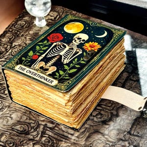 May include: A vintage-style journal with a cover depicting a skeleton, moon, and flowers. The cover reads "THE OVERTHINKER". It has a cream-coloured strap with metal hardware. The pages have a distressed, aged appearance.