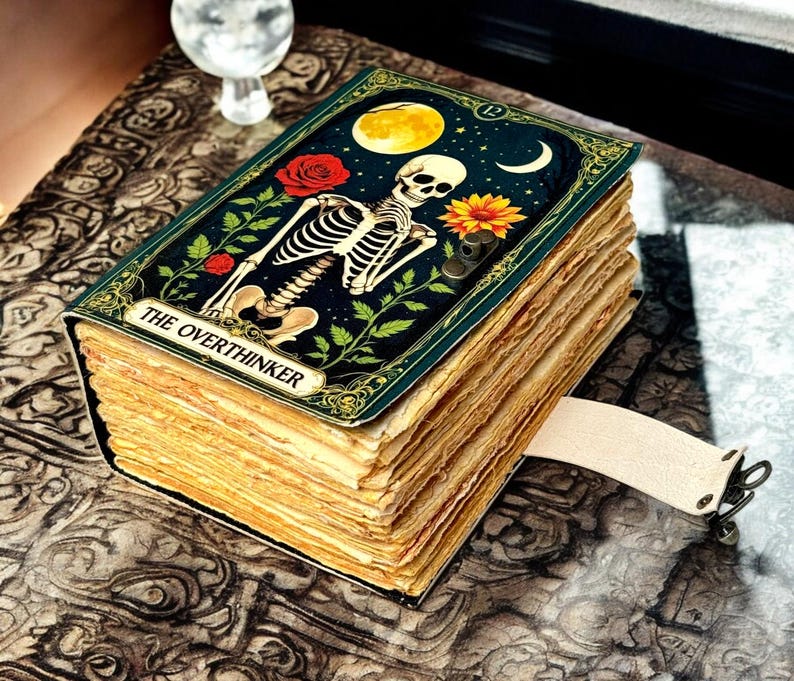 May include: A vintage-style journal with a cover depicting a skeleton, moon, and flowers. The cover reads "THE OVERTHINKER". It has a cream-coloured strap with metal hardware. The pages have a distressed, aged appearance.