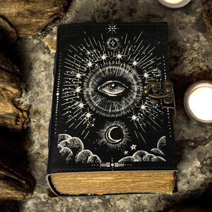 Puede incluir: Diario negro con dise&ntilde;o de ojo y celestial. La portada presenta una ilustraci&oacute;n detallada de un ojo, estrellas y una luna creciente. El diario tiene un cierre de metal y p&aacute;ginas envejecidas, lo que sugiere un tema vintage o m&iacute;stico.