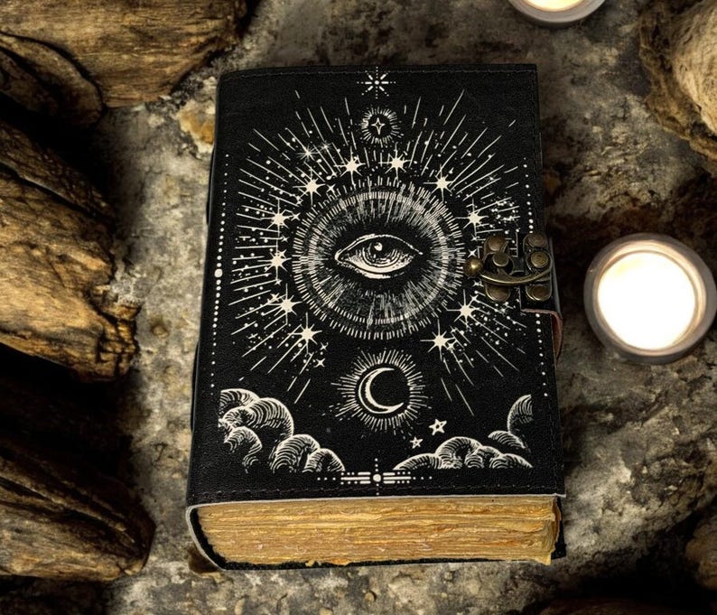Puede incluir: Diario negro con dise&ntilde;o de ojo y celestial. La portada presenta una ilustraci&oacute;n detallada de un ojo, estrellas y una luna creciente. El diario tiene un cierre de metal y p&aacute;ginas envejecidas, lo que sugiere un tema vintage o m&iacute;stico.
