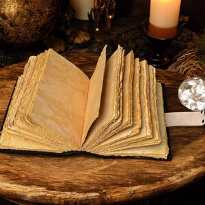 Puede incluir: Un libro antiguo abierto, con p&aacute;ginas amarillentas y envejecidas. El libro est&aacute; encuadernado en negro y descansa sobre una superficie de madera. Una bola de cristal y velas en el fondo crean un tema m&iacute;stico.