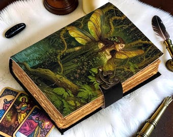 May include: A rectangular journal with a hard cover featuring a printed illustration of a winged fairy in a forest setting. The book has thick, deckled-edge pages and a black leather strap closure secured with a metallic latch on the front cover.