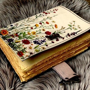May include: A handmade journal with a floral design on the cover. The cover features a cream background with colorful flowers and green leaves. The journal has aged, textured pages and a leather strap closure.