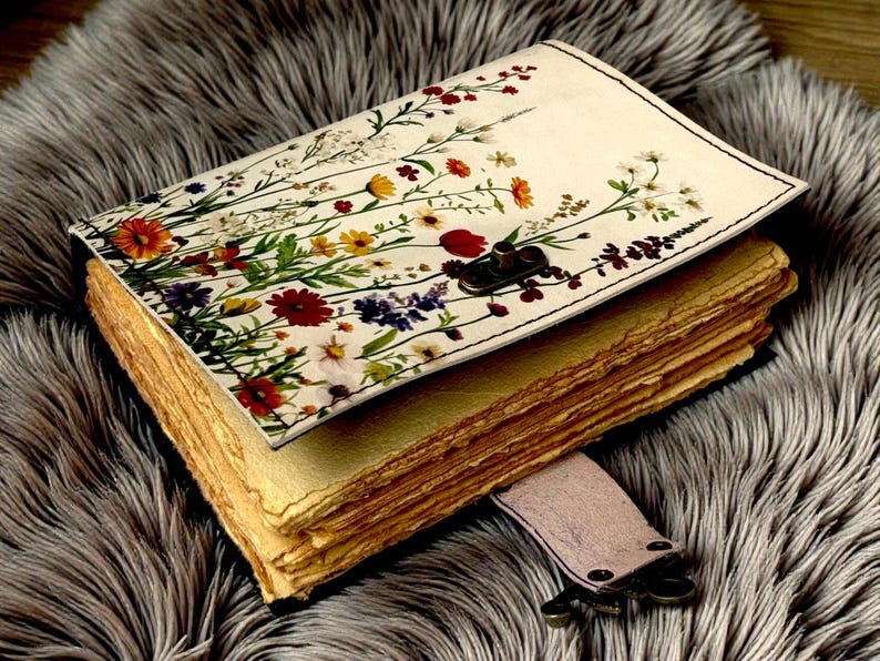 May include: A handmade journal with a floral design on the cover. The cover features a cream background with colorful flowers and green leaves. The journal has aged, textured pages and a leather strap closure.