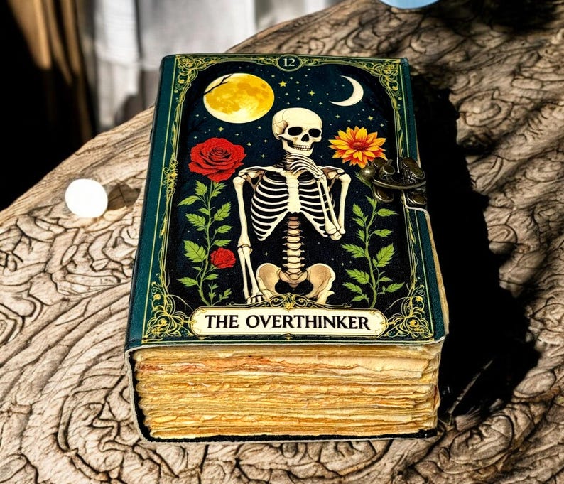 May include: A decorative book with a dark teal cover featuring a skeleton illustration under a moon and stars. The book is titled "THE OVERTHINKER" and has a clasp closure. The pages are aged and yellowed.