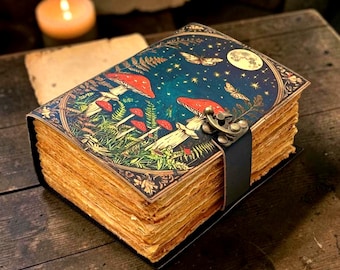 May include: A rectangular journal with thick, deckled-edge paper pages. The cover features a dark blue night sky illustration with red mushrooms, green ferns, stars, a moon, and butterflies. A black leather strap with a brass-toned metal latch closure.