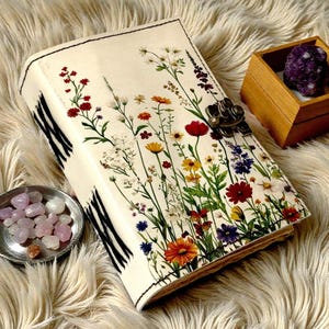 May include: A cream-colored journal with a floral design featuring red, yellow, and purple flowers. The journal has a metal clasp and black and white striped edges. A small wooden box with a purple stone and a small silver dish with pink stones are also present.