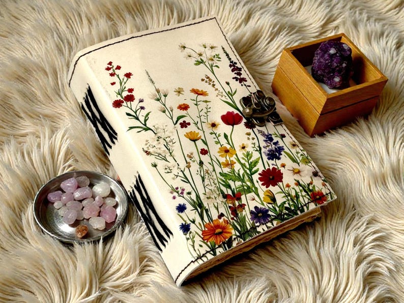 May include: A cream-colored journal with a floral design featuring red, yellow, and purple flowers. The journal has a metal clasp and black and white striped edges. A small wooden box with a purple stone and a small silver dish with pink stones are also present.