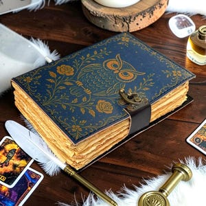 May include: A rectangular journal with a dark blue cover featuring a gold-toned owl and floral pattern. The book has thick, deckled-edge pages and is secured with a dark brown leather strap and a metallic latch closure on the front edge.