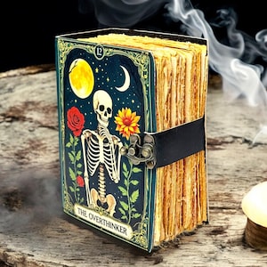 May include: A decorative journal with a dark teal cover featuring a skeleton illustration, a full moon, and a crescent moon. The journal is bound with a black strap and clasp. The text "THE OVERTHINKER" is visible on the cover.