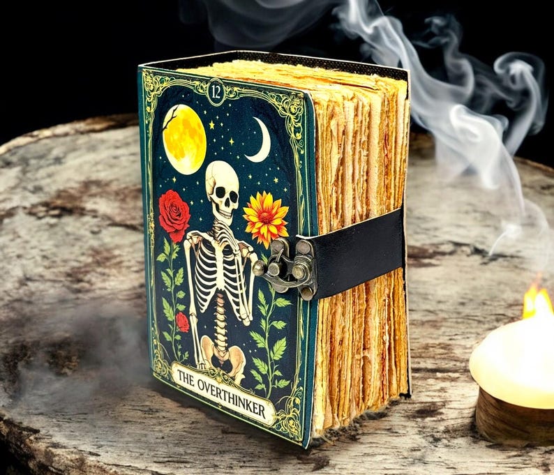 May include: A decorative journal with a dark teal cover featuring a skeleton illustration, a full moon, and a crescent moon. The journal is bound with a black strap and clasp. The text "THE OVERTHINKER" is visible on the cover.