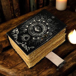 Puede incluir: Un diario negro con un dise&ntilde;o de ojo y celestial en la portada. Las p&aacute;ginas est&aacute;n envejecidas y se inserta un marcap&aacute;ginas de cinta beige. El diario est&aacute; sobre una superficie de madera, con velas en el fondo.