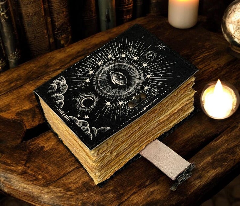 Puede incluir: Un diario negro con un dise&ntilde;o de ojo y celestial en la portada. Las p&aacute;ginas est&aacute;n envejecidas y se inserta un marcap&aacute;ginas de cinta beige. El diario est&aacute; sobre una superficie de madera, con velas en el fondo.