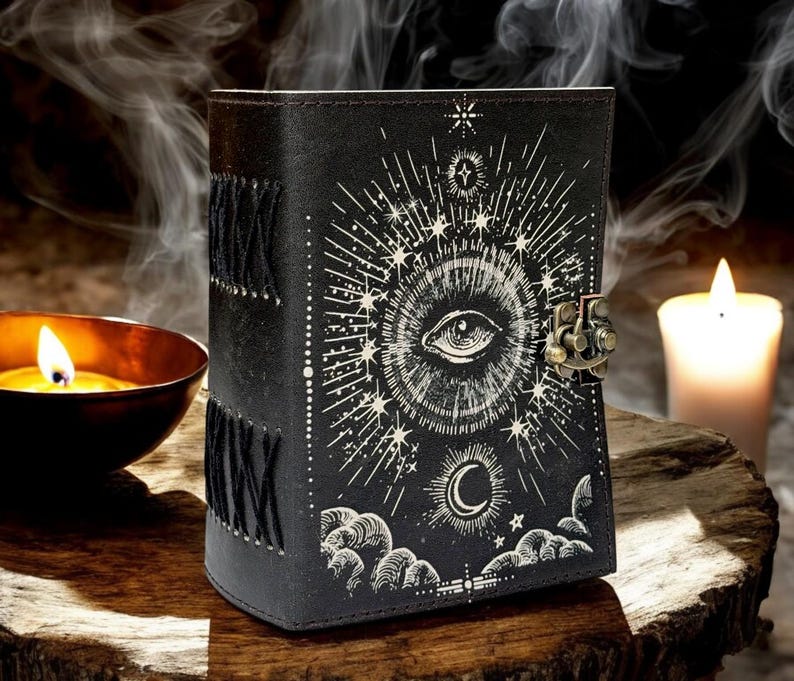 Puede incluir: Diario de cuero negro con un dise&ntilde;o celestial. La portada presenta un s&iacute;mbolo de ojo central rodeado de l&iacute;neas radiantes, estrellas y una luna creciente. El diario tiene un cierre de metal y costuras decorativas a lo largo del lomo.